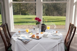 Mesa de café da manhã posta com croissants, suco de laranja e um vaso de rosas vermelhas, com vista para um gramado verde.