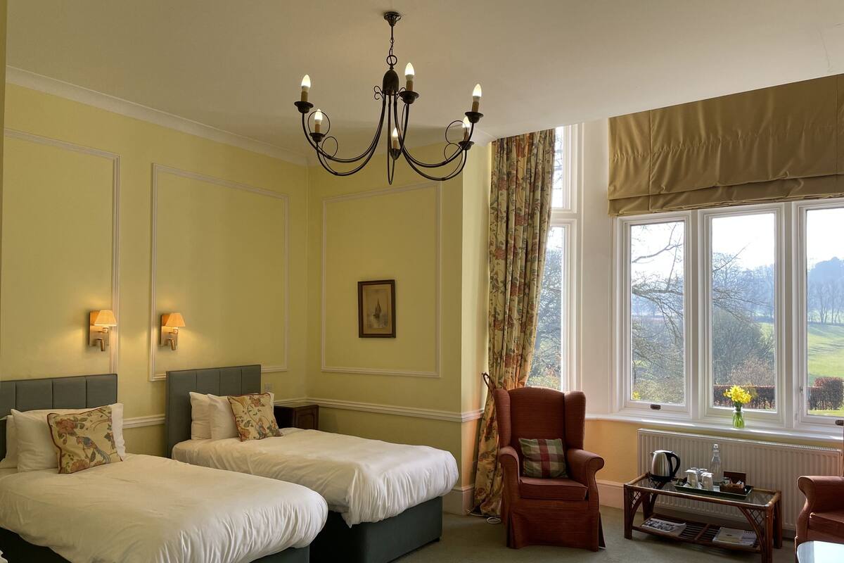 A hotel room with two beds, a sitting area with a chair, a table, and a view of the outdoors through large windows.