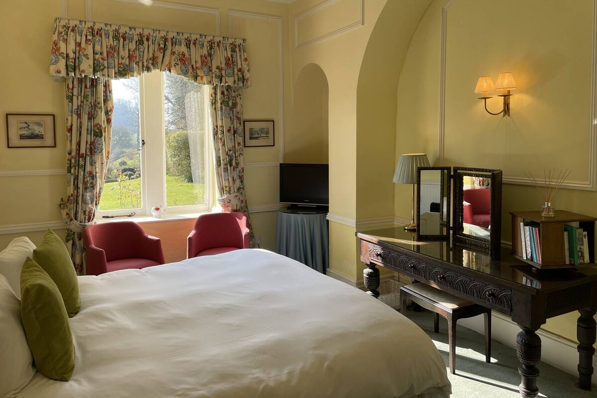 A hotel room with a large bed, a desk with a mirror, a television, and a view of the garden through the window.
