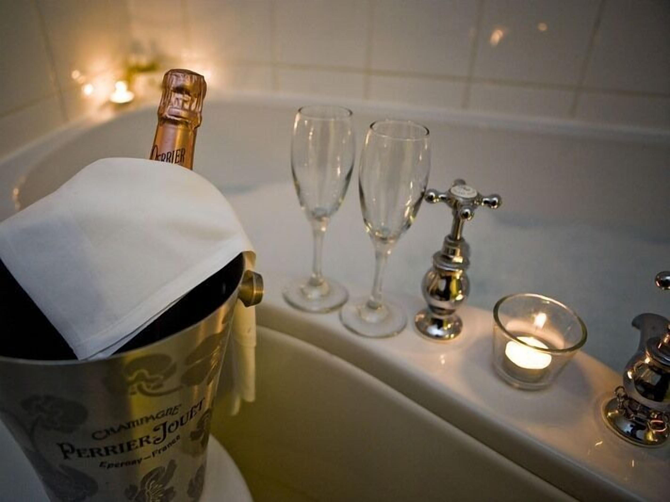 A champagne bottle in a bucket, two glasses, and a bathtub with a candle.