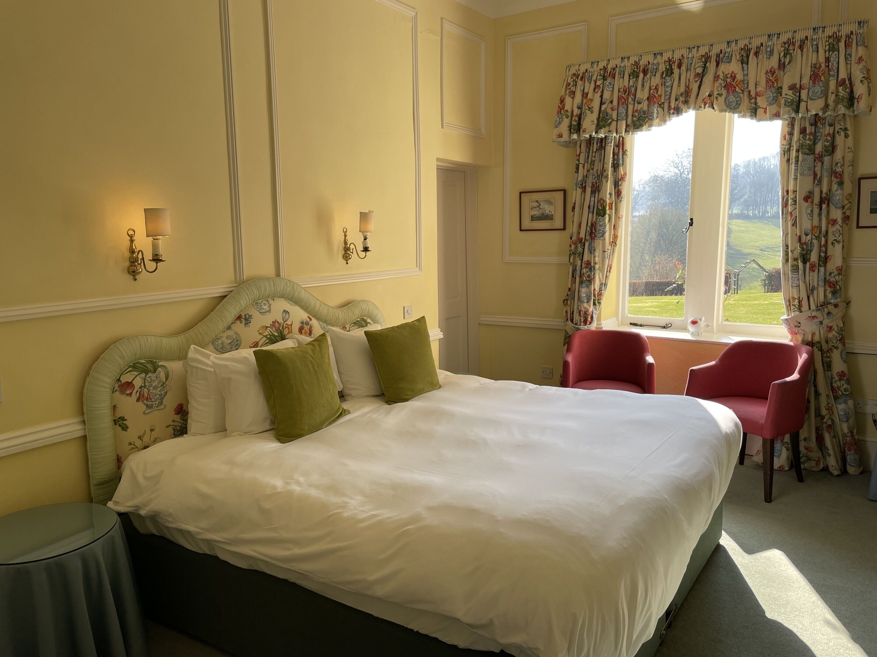 Un lit bien fait, avec du linge de lit blanc et des coussins verts, une chaise rouge et une fenêtre ornée de rideaux à motifs floraux.
