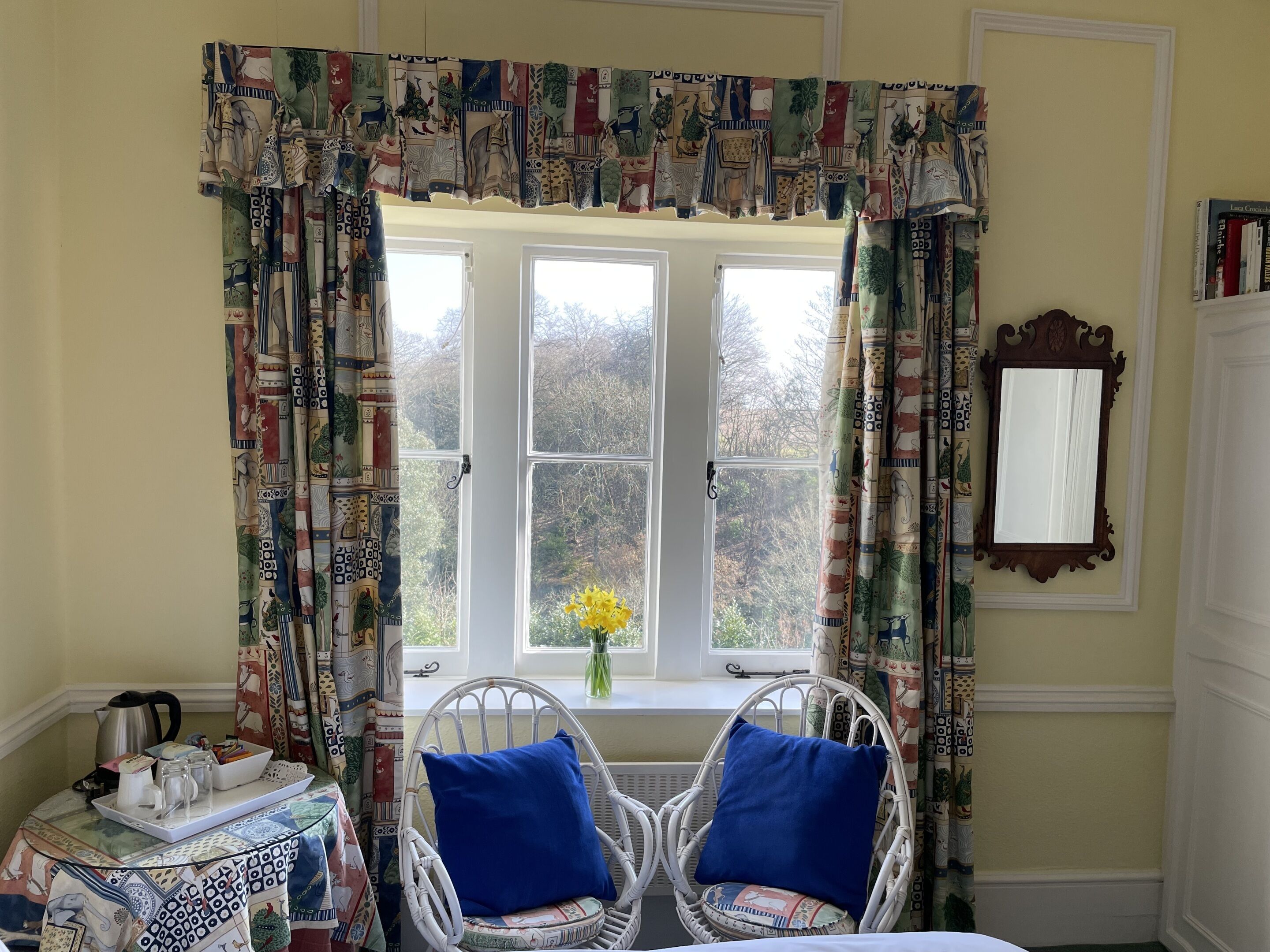 Een kamer met een groot raam, twee rieten stoelen met blauwe kussens, een bijzettafeltje met een dienblad en een spiegel aan de muur.