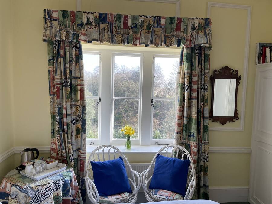 Een kamer met een groot raam, twee rieten stoelen met blauwe kussens, een bijzettafeltje met een dienblad en een spiegel aan de muur.