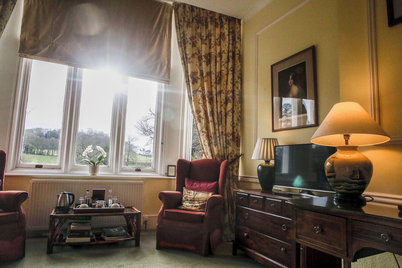 Um quarto com uma janela grande, uma poltrona vermelha, uma cômoda de madeira, uma mesa com abajur e um quadro na parede.