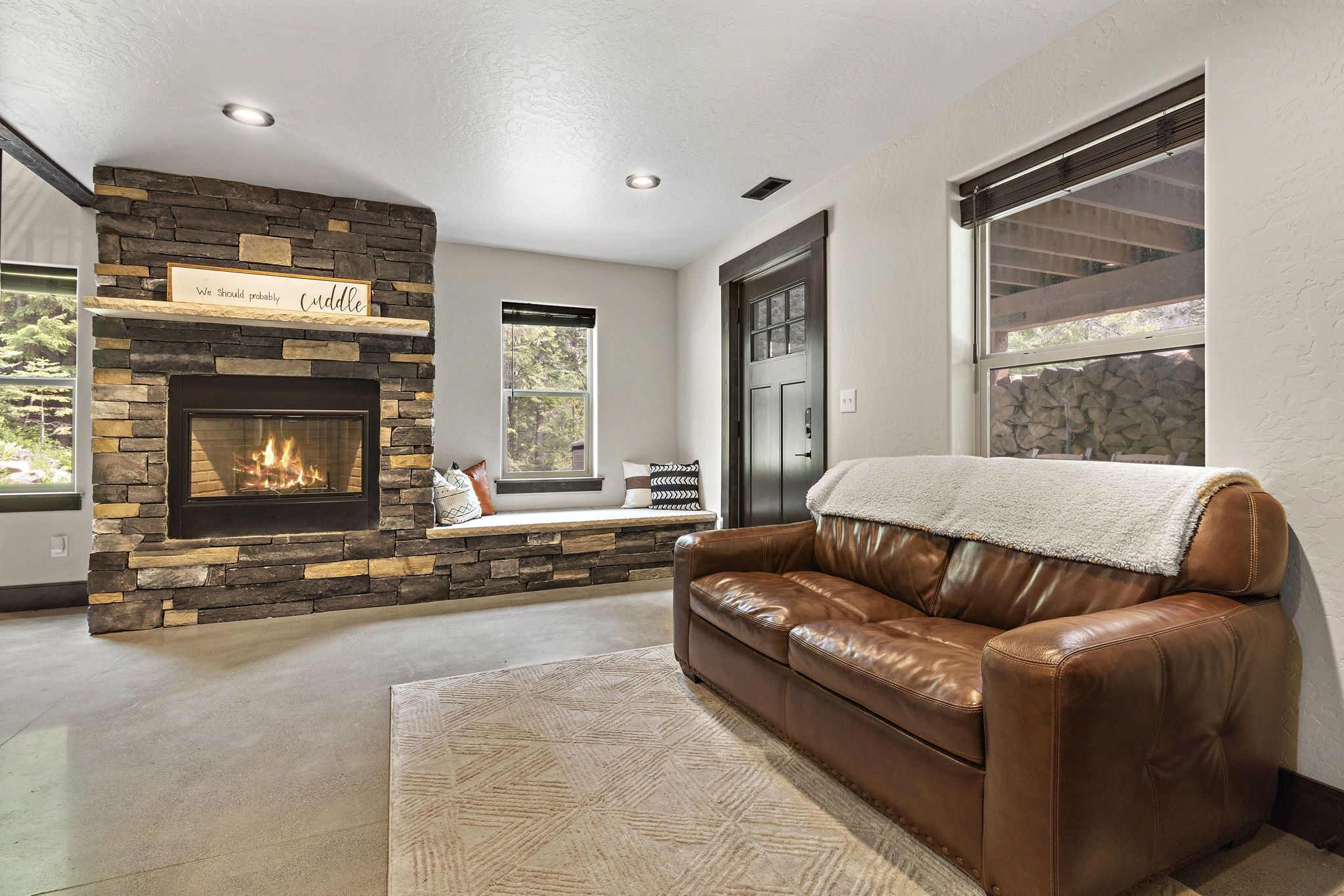 A living room with a stone fireplace, a leather sofa, and a window with a view of trees.