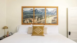 A hotel room with a large bed, white bedding, a mountain landscape painting above the bed, and a nightstand with a lamp and a clock. - Historic Flat in Downtown Boise (Boise)