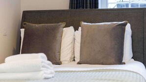 Ein ordentlich bezogenes Bett mit Kissen und einem gefalteten Handtuch, ein Kopfteil und ein Fenster mit Vorhängen.