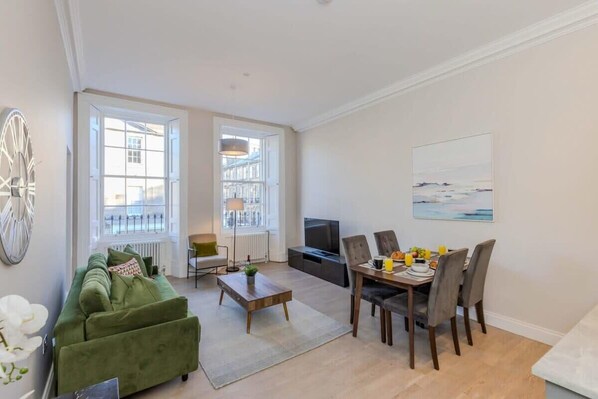 A modern living room with a green sofa, dining table, and chairs, a TV, and large windows. - Edinburgh City Centre 2 Bed Apartment (Edinburgh)