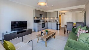 Een moderne woonkamer met een flatscreen televisie, een houten salontafel, groene banken en een keuken met grijze keukenkastjes.