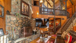 A cozy cabin interior with a stone fireplace, wooden walls, a television, and a staircase leading to an upper level. - Rustic Beauty in Chamberlands Full Beach & Pool (Homewood)