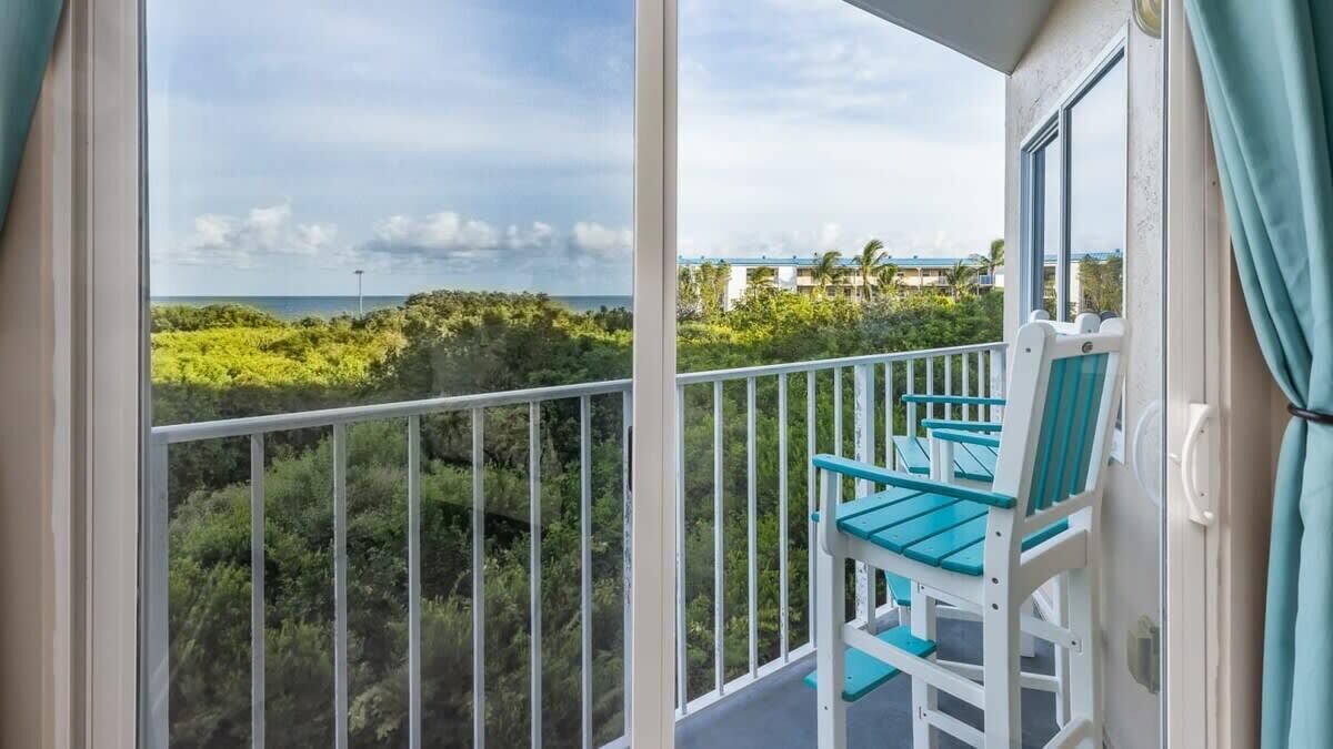 A balcony with a view of the ocean and vegetation, a white railing, and a blue chair.