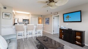 A modern living room with a sofa, a coffee table, and a TV. There is a kitchen area with a microwave and a dining set.