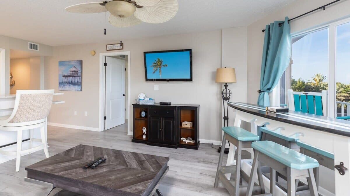 A modern living room with a television, a coffee table, a seating area with bar stools, and a view of the outdoors.