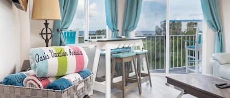 A cozy room with a bed, a table, and a chair. There's a view of the outdoors through large windows.