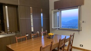 A dining area with a wooden table and chairs, a fruit bowl, and a window with a view of the mountains.