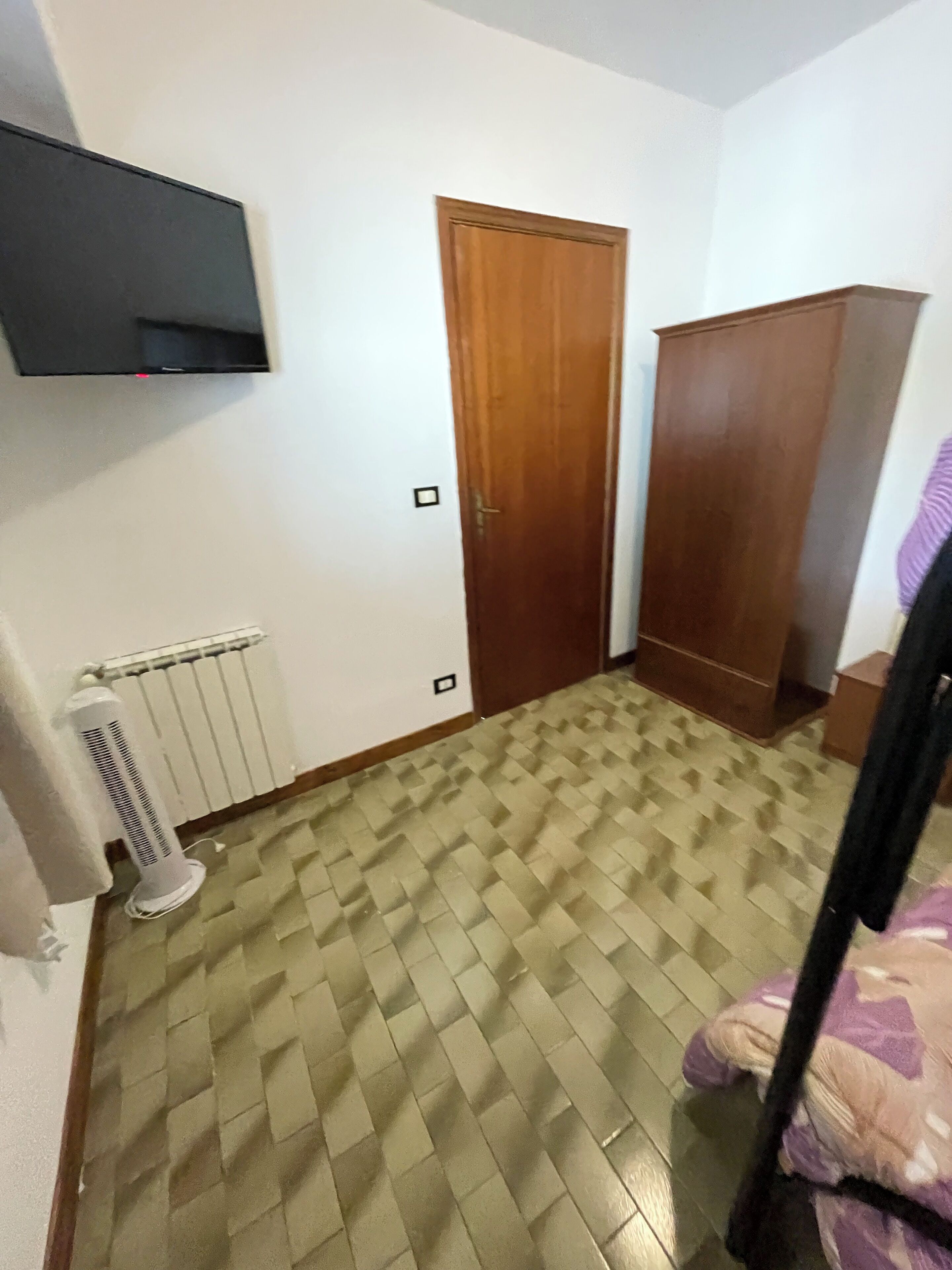 Ein Zimmer mit einem gemusterten Boden, einer Holztür, einem Kleiderschrank, einem Heizkörper und einem an der Wand befestigten Fernseher.