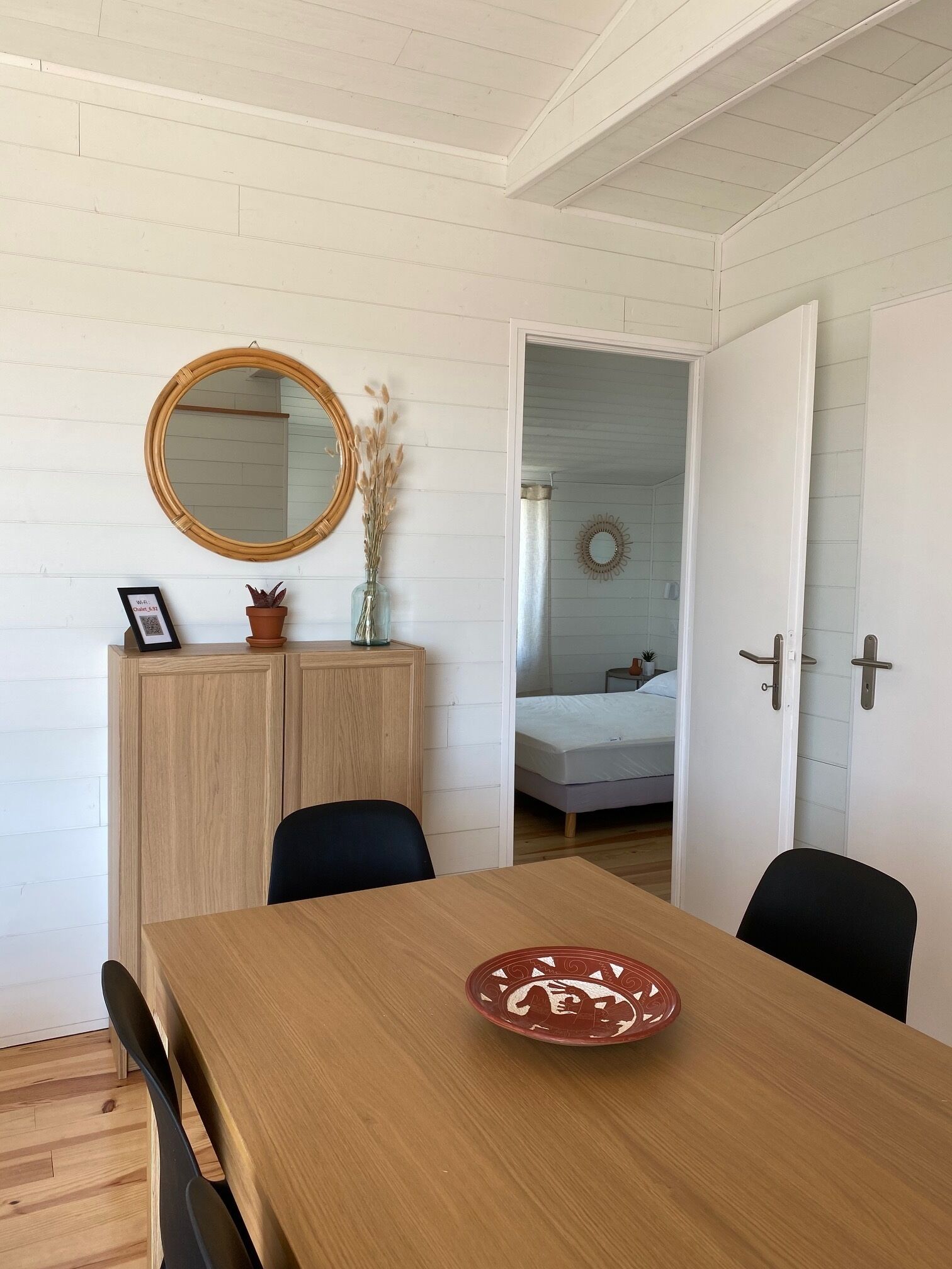 A room with a wooden dining table, chairs, a mirror, a cabinet, and a bed in the background.