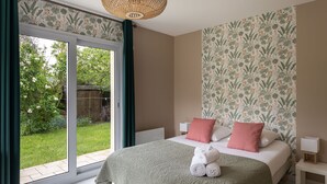 A bedroom with a large sliding glass door leading to a garden, a bed with a green blanket and pink pillows, and a wall with a floral pattern.