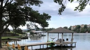 A lakeside area with a sandy beach, a covered seating area with chairs, and a dock extending into the water. - Lake house with access to the best beach for swimming & fishing on Lake Hamilton (Hot Springs)