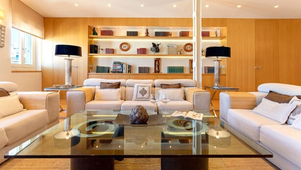Un salon moderne avec une table basse en verre, des canapés blancs et une bibliothèque en bois contenant des livres et des objets décoratifs.