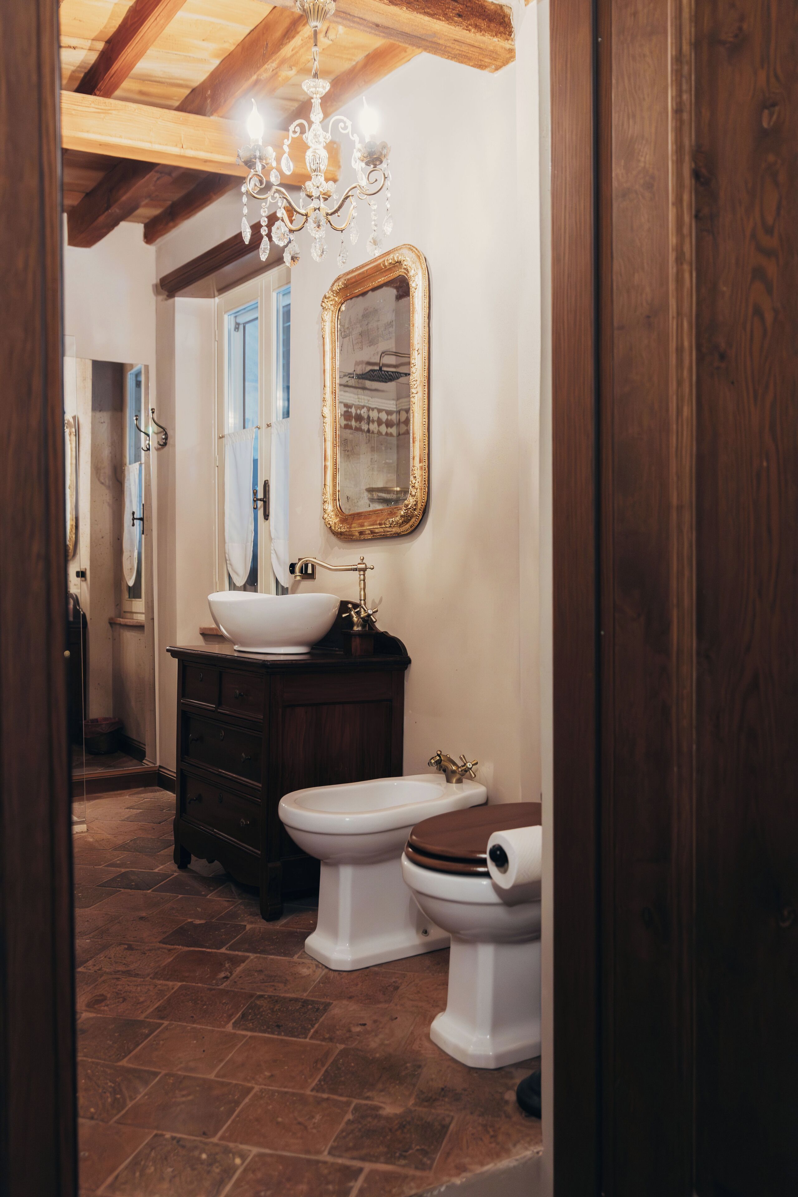 Een badkamer met een houten vloer, een witte wastafel, een bidet en een spiegel met gouden lijst.