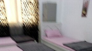 Ein Zimmer mit zwei Betten, eines mit rosa Matratze und das andere mit schwarzer Matratze. Eine weiße Wand mit Fenster und einem kleinen Bilderrahmen.