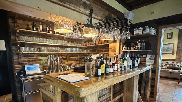 Una zona de bar rústica con estantes de madera, una barra alargada y suelo de baldosas.