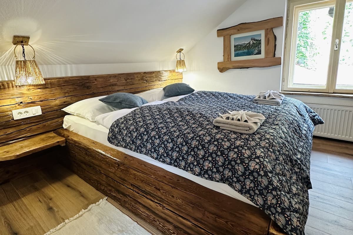 A bedroom with a wooden headboard, a large bed with a patterned bedspread, two bedside lamps, a window with a view of greenery, and a radiator.