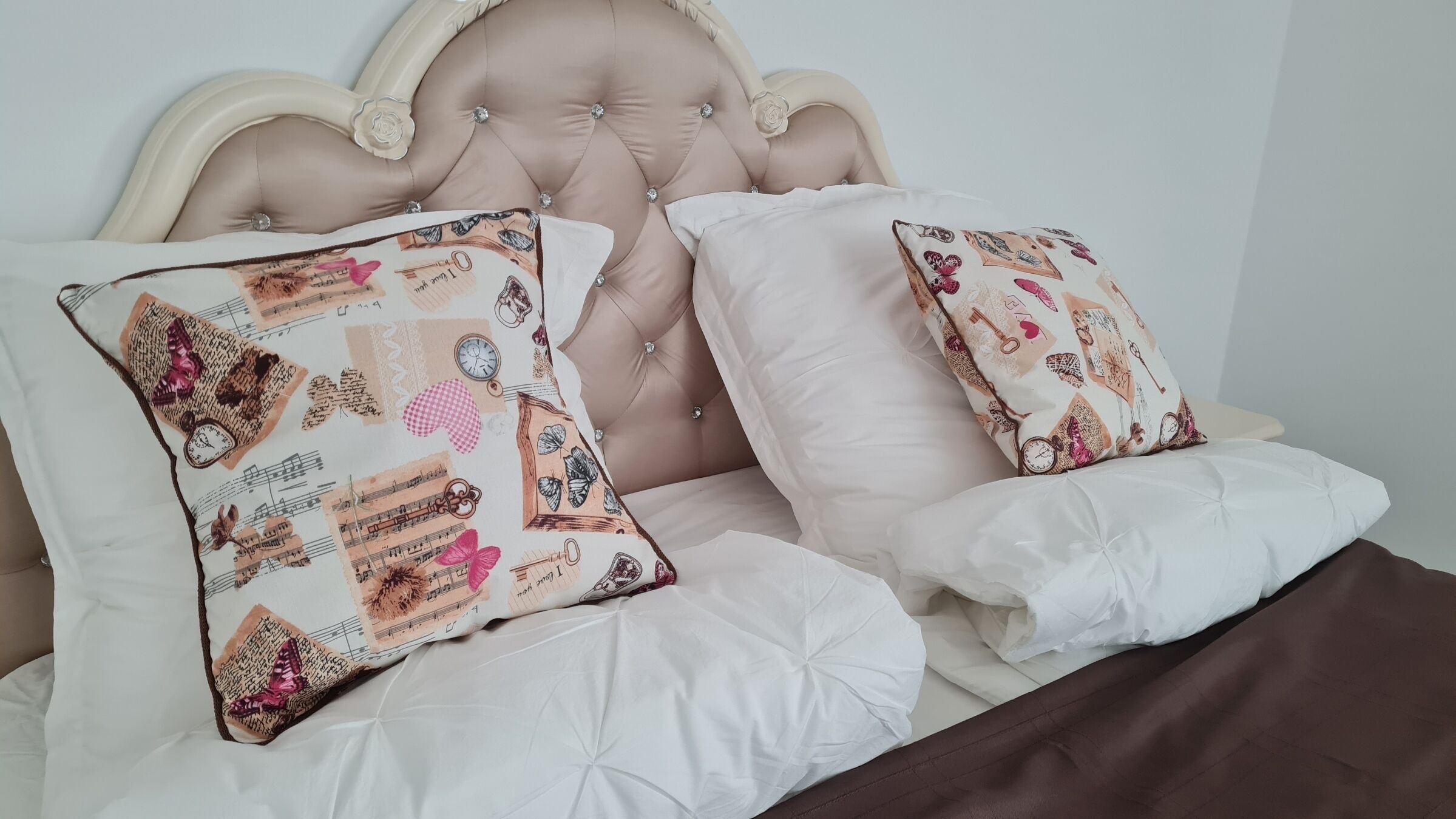 A bed with a tufted headboard, two decorative pillows with a vintage design, and a white comforter.