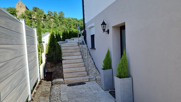 A narrow pathway leading to a modern building with a stone staircase, a white wall with a black lantern, and a stone tower in the background.