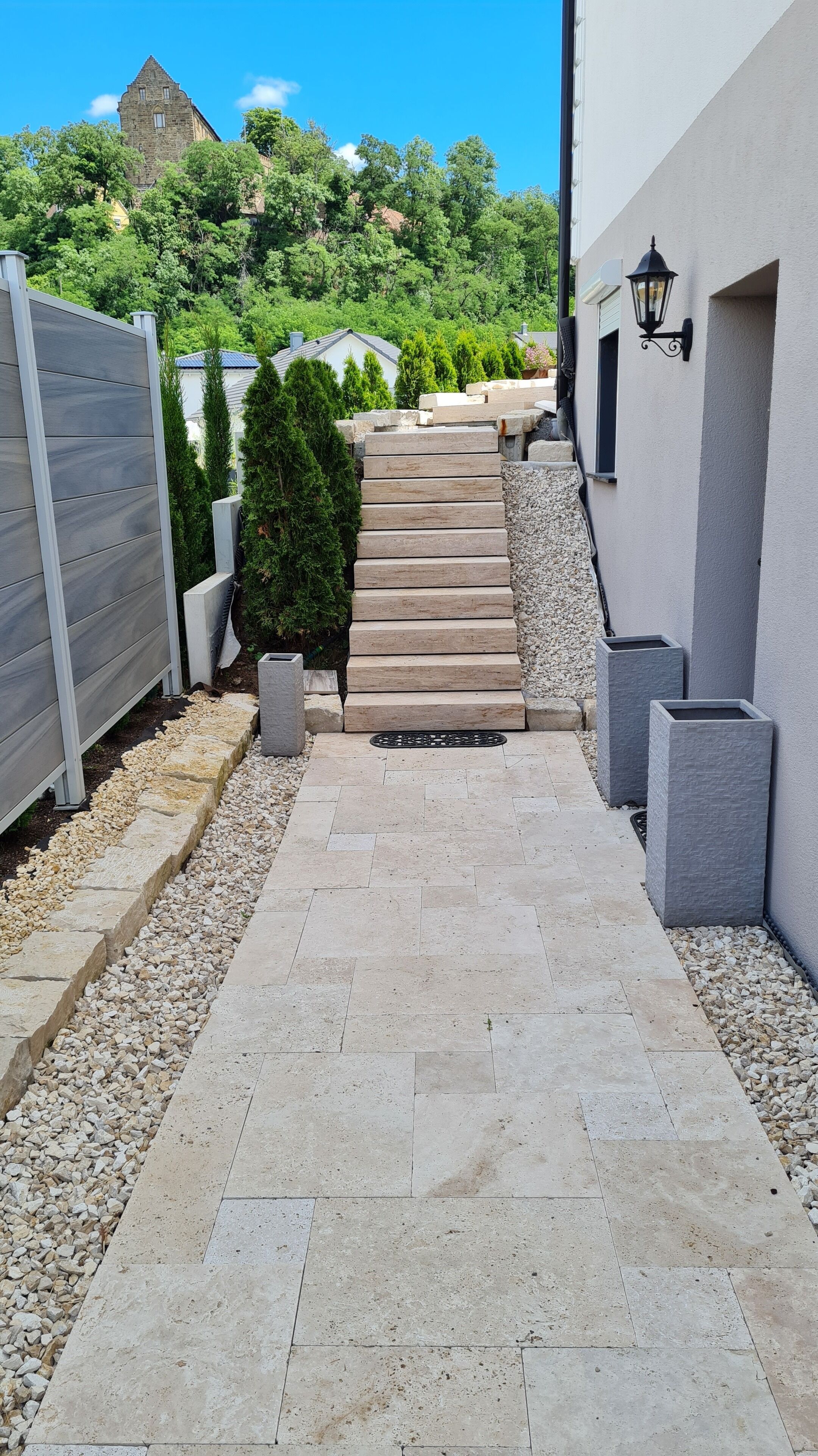 A paved pathway leading to a building with a stone staircase, surrounded by gravel and small trees.
