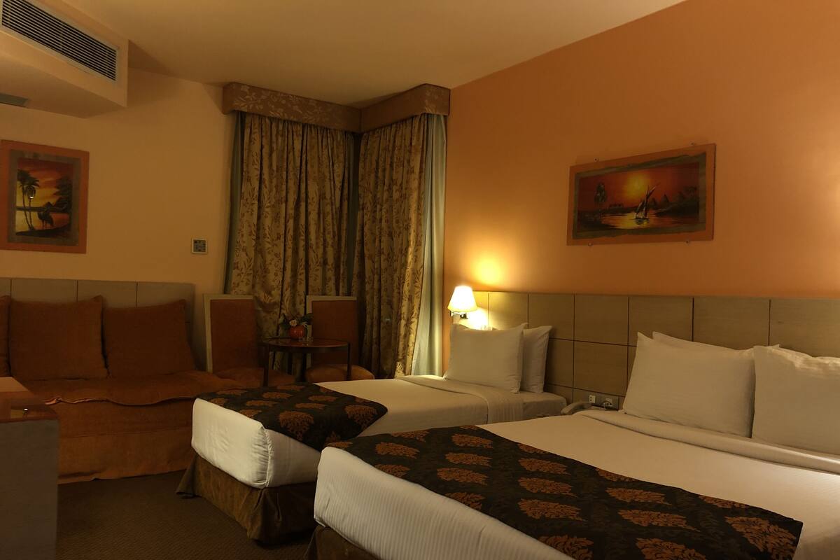 A hotel room with two beds, a sofa, a table with a lamp, and two paintings on the walls.