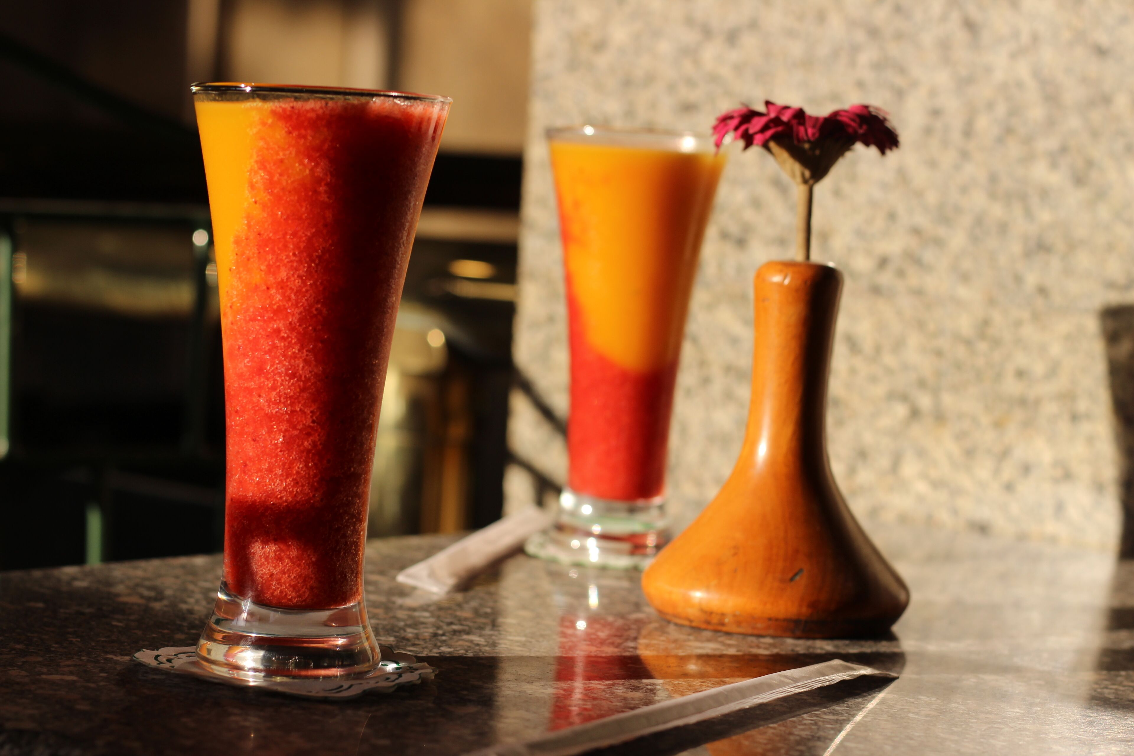 Deux grands verres contenant une boisson de couleur rouge-orangée sont posés sur le comptoir d’un bar, et un vase en bois avec une fleur séchée se trouve à l’arrière-plan.