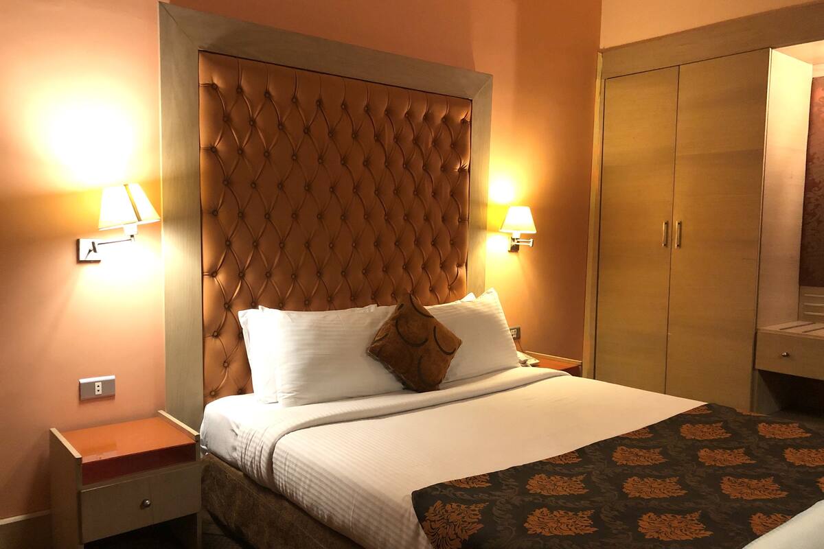 A hotel room with a tufted headboard, a bed with white linens and a brown throw, a nightstand with a lamp, and a wardrobe.