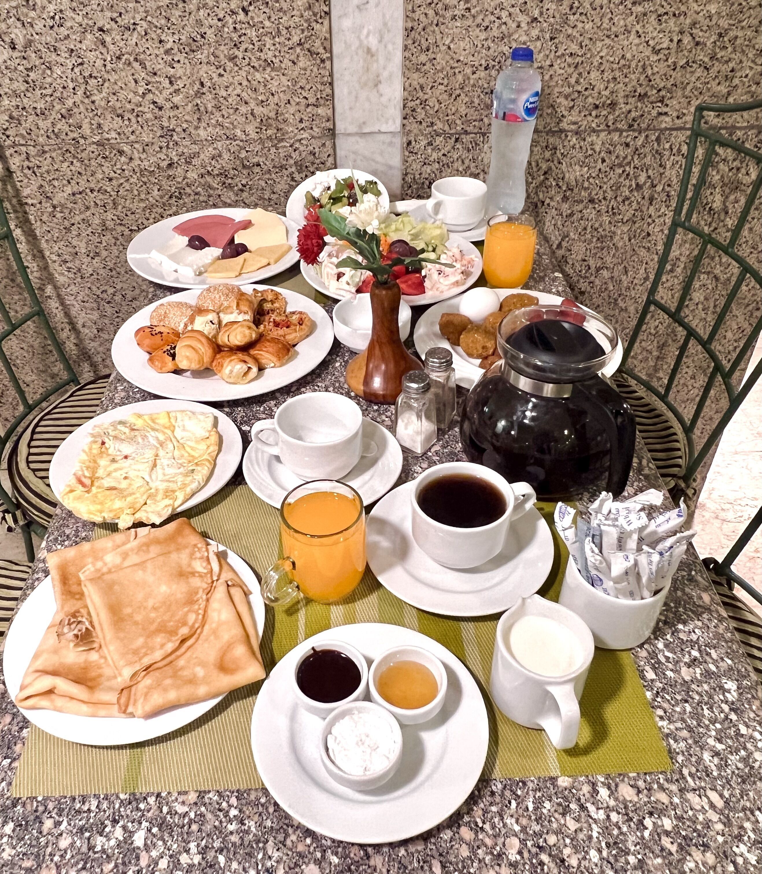 A breakfast buffet with various pastries, fruits, and beverages on a table with a green chair and a vase of flowers.