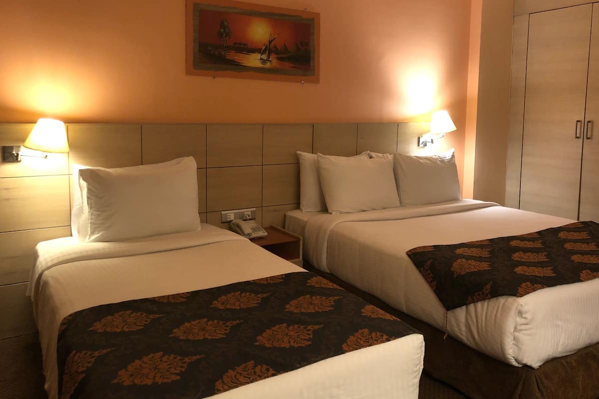 A hotel room with two beds, each with white linens and a floral-patterned throw. There are two bedside lamps, a painting on the wall, and a wardrobe with a mirror.