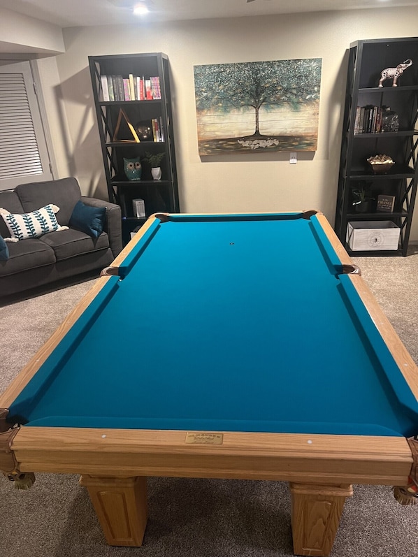 A pool table in a room with a couch, bookshelves, and a painting on the wall.