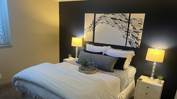Un letto rifatto con un comofort bianco e cuscini, due comodini con lampade, un quadro e una finestra con persiane.