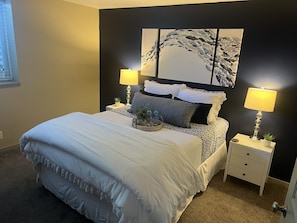 A neatly made bed with a white comforter and pillows, two bedside tables with lamps, a wall art piece, and a window with blinds.