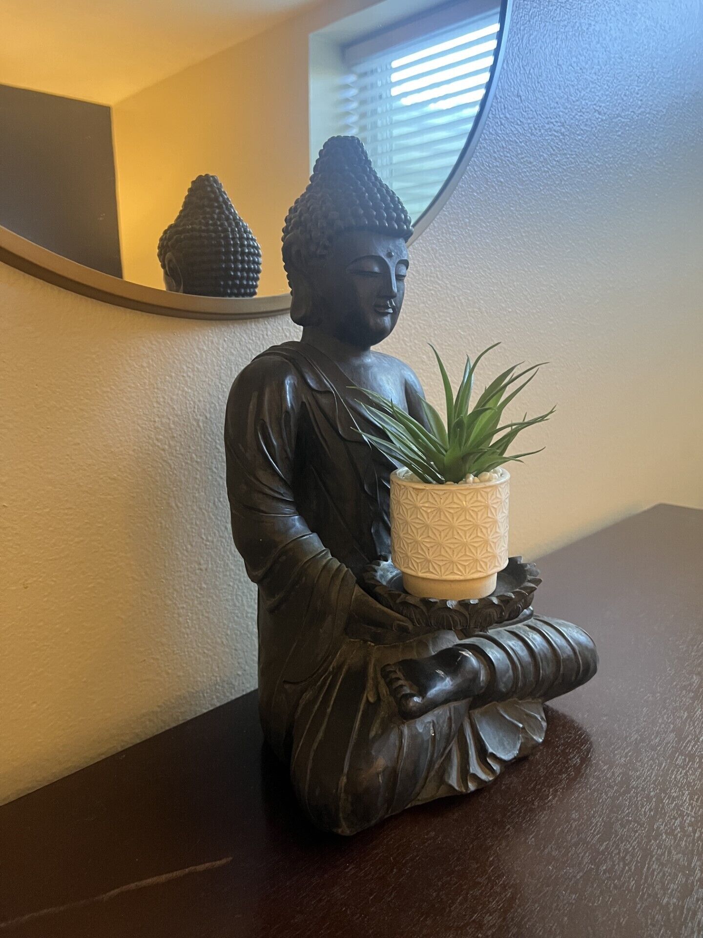 A statue of a seated figure holding a plant, placed on a dark surface with a mirror reflecting part of the room.