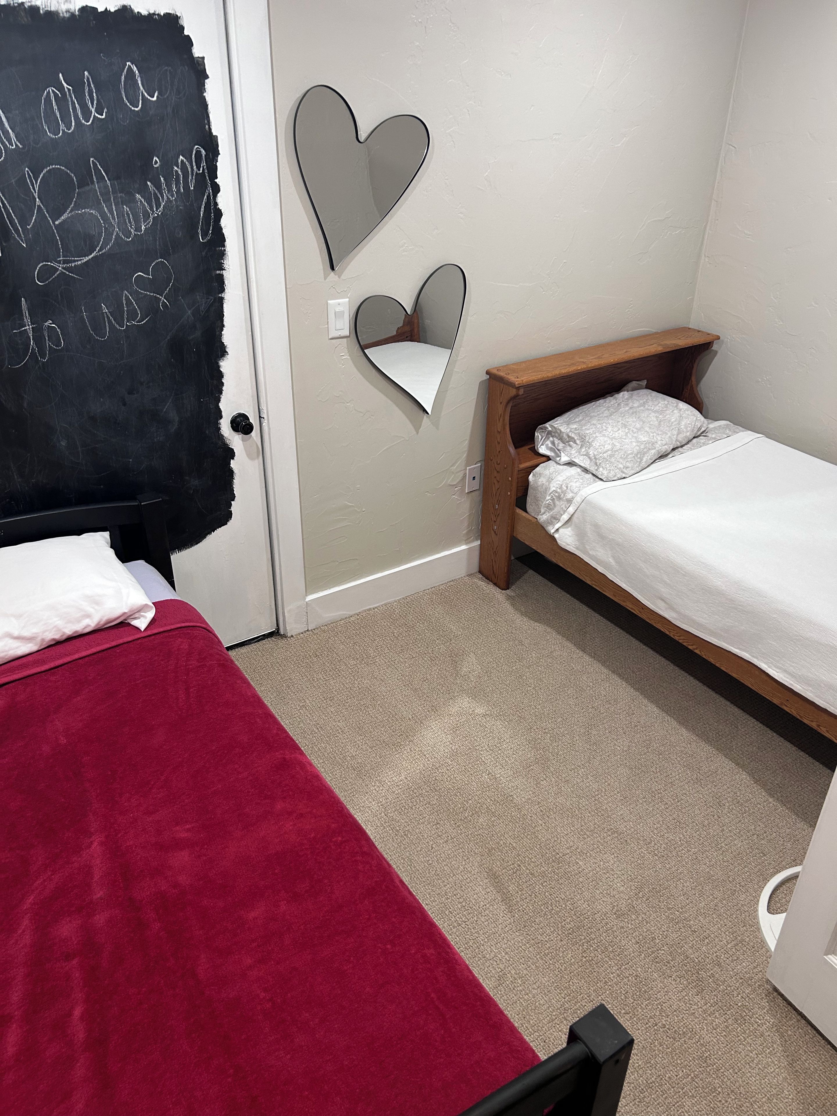 Una camera da letto con due letti, uno con copriletto rosso e l'altro con copriletto bianco. Sono presenti specchi a forma di cuore sulla parete e una lavagna con scritte.
