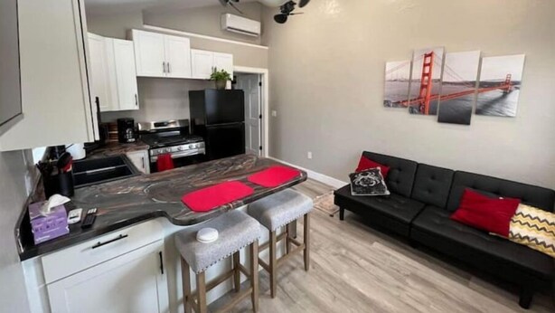 Et moderne køkken med hvide skabe, et sort køleskab og en marmor bordplade. I opholdsstuen er der en sort sofa med røde og gule puder.