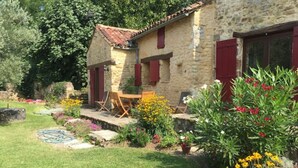 Une maison en pierre avec un toit en tuiles, un petit patio avec une table et des chaises, le tout entouré d’un jardin bien entretenu où poussent différentes fleurs et arbustes.