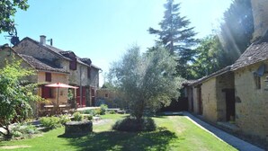Une cour bordée de bâtiments en pierre, avec un petit arbre et une table parasol rouge.