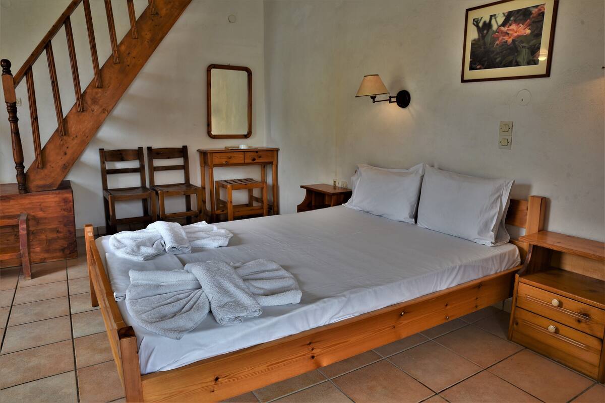 A neatly made bed with white linens and rolled towels, a wooden nightstand, a wooden chair, a small wooden table, a wall-mounted lamp, and a wooden staircase.