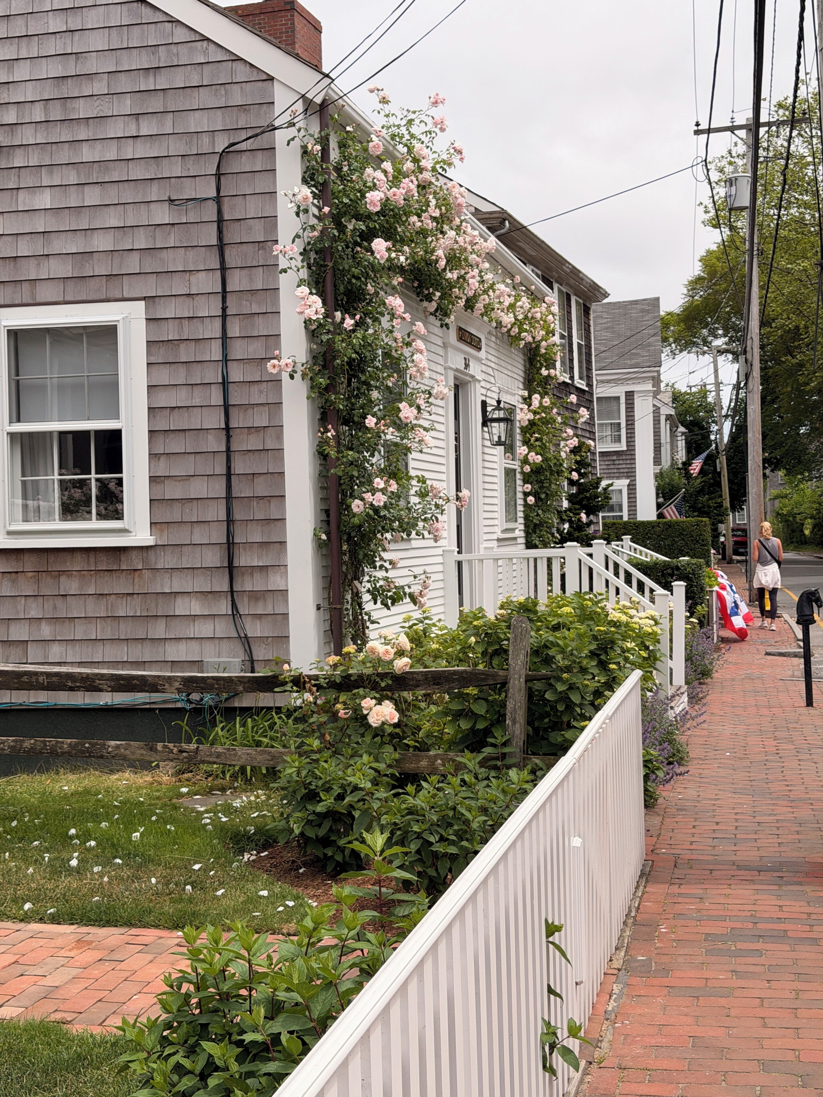 A residential house with a white picket fence, a garden with pink flowers, and a brick pathway.