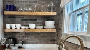 Una cocina con pared de ladrillo, estantes de madera, pileta de acero inoxidable y una ventana con cortinas.