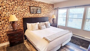 Een slaapkamer met een bed, een wand van houten balken, twee nachtkastjes met lampen, en een raam met jaloezieën.
