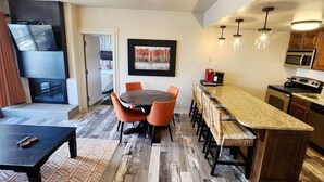 Een moderne keuken met een centraal eiland, een eethoek met oranje stoelen, en een aan de muur bevestigde televisie.
