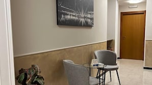 A hallway with a potted plant, two grey chairs, a small table, and a black and white painting on the wall. - Shelter Affittacamere (Genoa)